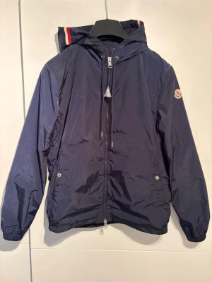 Mörkblå vindjacka från Moncler - Säljer en mörkblå Moncler vindjacka i väldigt fint skick. Stilren och lätt jacka som passar perfekt till vardags och kan enkelt matchas med olika outfits.  tagsen från Moncler sitter inte kvar på jackan men medföljer. Ingen förpackning, påse eller annat tillbehör medföljer – endast jackan och korten/tagsen. Storlek: 2 (M)  📦 Endast frakt.