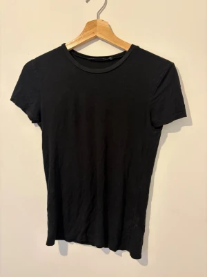 Svart basic t-shirt med rund hals - En enkel svart t-shirt med rund hals och korta ärmar. Modellen är rak och klassisk, perfekt för att matcha med olika stilar. Materialet ser ut att vara mjukt och bekvämt, troligen bomull eller bomullsblandning.