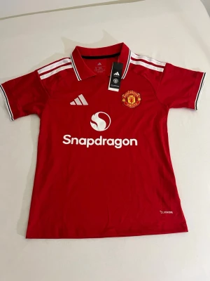 Manchester United röd matchtröja - Snygg röd Manchester United matchtröja från Adidas med vita detaljer och klubbmärke på bröstet. Tröjan har vit logga och sponsortryck, samt klassiska Adidas-ränder på axlarna. Tillverkad i lätt och ventilerande material.