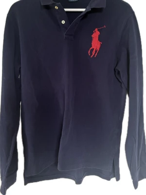 Ralph Lauren  långärmad pikéskjorta - Mörkblå långärmad pikéskjorta från Ralph Lauren med klassisk krage och knappar framtill. Stor röd broderad logga på bröstet och röd detalj på ärmen. Skjortan har en avslappnad passform och är tillverkad i bomull.