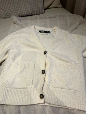 Vit kofta från Vero Moda - Mysig vit kofta från Vero Moda med stora knappar framtill och två rymliga fickor. Koftan har ribbade muddar och en mjuk, fluffig känsla som gör den perfekt för kyliga dagar. Enkel och stilren design som passar till det mesta.