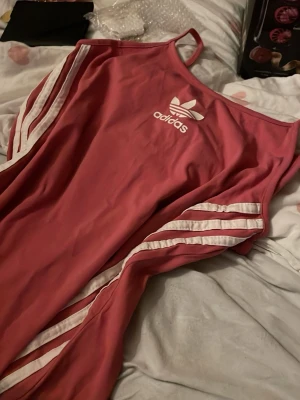 Rosa linnetopp från Adidas - Snygg rosa linnetopp från Adidas med vita ränder längs sidorna och Adidas-logga framtill. Tunt material och smala axelband ger en sportig och trendig look. Perfekt för dig som gillar streetstyle och sportiga vibes.