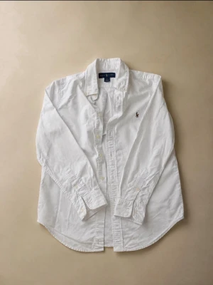 Vit skjorta från Ralph Lauren - Klassisk vit skjorta från Ralph Lauren med knappstängning och broderad logga på bröstet. Tillverkad i bomull och har långärmad design med krage och manschetter. Perfekt för en clean och stilren look.