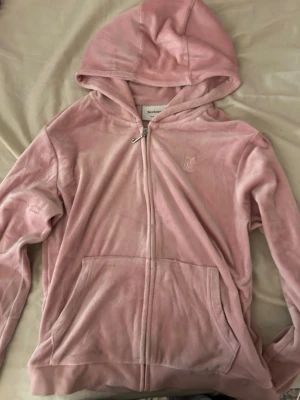 Ljusrosa velour hoodie med dragkedja - Mysig ljusrosa hoodie i mjuk velour med dragkedja framtill och stor ficka. Har huva och diskret broderad logga på bröstet. Perfekt för en chill och avslappnad stil.