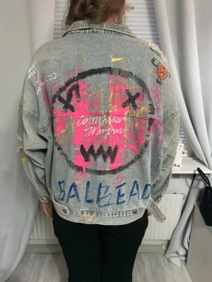 Oversized jeansjacka med graffiti - Unik oversized jeansjacka med graffiti-inspirerade målningar i rosa, blått och svart. Jackan har slitna detaljer, trasiga ärmar och handmålade texter och motiv på både rygg och ärm. Perfekt för dig som vill sticka ut med en streetstyle-look.
