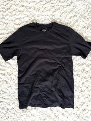 Svart basic t-shirt i bomull - En enkel svart t-shirt i mjuk bomull med rund hals och korta ärmar. Perfekt till alla outfits och har en klassisk passform som funkar till det mesta. Stilren och tidlös design utan tryck eller detaljer.