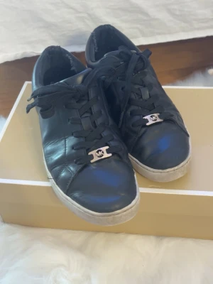 Svarta Michael Kors sneakers i läder - Snygga svarta sneakers från Michael Kors i äkta läder med vita sulor och klassisk låg design. Skorna har svarta snören och en metallbricka med MK-logga framtill. Perfekta för en stilren och trendig look.