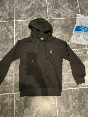 Svart hoodie från Polo Ralph Lauren - Snygg svart hoodie från Polo Ralph Lauren med klassisk huva och dragsnören. Har en broderad vit logga på bröstet och en stor magficka framtill. Tillverkad i mjukt material som känns skönt mot huden. Perfekt för en avslappnad och stilren look.