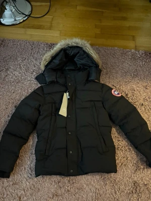 Svart dunjacka med pälskantad huva - Säljer en svart dunjacka från Canada Goose med pälskantad huva och klassisk logga på ärmen. Jackan har dragkedja och knappar framtill, två stora fickor och en varm, vadderad insida. Perfekt för kalla vinterdagar.