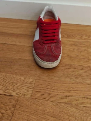 Röda låga sneakers med vita detaljer - Snygga röda sneakers med vita detaljer och röda skosnören. Skon har en klassisk låg siluett och en vit sula. Materialet ser ut att vara mocka eller liknande tyg. Perfekt för dig som vill sticka ut med färg på fötterna. Priset går att pruta då jag ska köpa ljud till bilen, så måste bli av med den snabbt😀