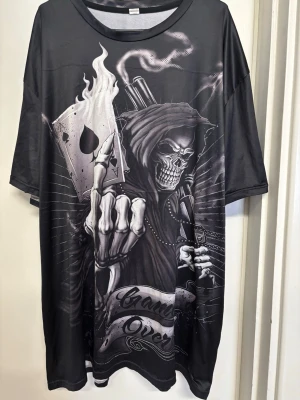 Svart oversized t-shirt med dödskalletryck - Cool svart oversized t-shirt med stort tryck av en dödskalle i hoodie, spelkort och rökdetaljer. T-shirten har korta ärmar och rund hals. Perfekt för dig som gillar edgy och unika motiv.någon defekt se bild