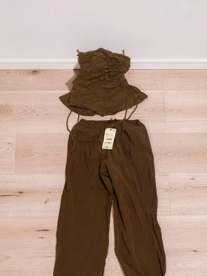Brunt set med topp och byxor från Zara - Säljer ett brunt tvådelat set med en bandeau topp med rynkade detaljer och matchande vida byxor med resår i midjan. Setet är gjort i ett mjukt, tunt tyg som ger en avslappnad look. Perfekt för dig som gillar trendiga och bekväma plagg. Säg till om ni vill ha bättre bilder :)