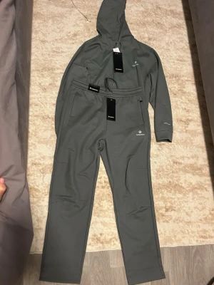 Grått loungewear-set från Salomon - Mysigt grått loungewear-set från Salomon med huva och dragsko på tröjan samt matchande byxor med resår i midjan. Båda delarna har Salomon-logga framtill och mjukt material som passar perfekt för chill hemma. Snygg minimalistisk design och praktiska fickor.