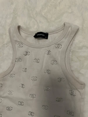 Vit ribbad linnetopp  - Supersnygg vit ribbad linnetopp med glittriga detaljer i form av små loggor över hela toppen. Ärmlös modell med rund halsringning och stretchigt material som sitter bekvämt. Perfekt för dig som vill ha en lyxig och trendig look. Storlek S