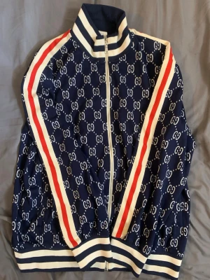 Blå zip-jacka med GG-mönster cardigan  - Snygg blå zip-jacka med vitt GG-mönster över hela plagget. Jackan har vita och röda ränder längs ärmarna samt ribbade muddar och krage i blått och vitt. Perfekt för dig som gillar sportig och trendig stil.