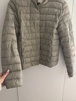 Ljusbeige pufferjacka med tryckknappar - Säljer en stilren ljusbeige pufferjacka med raka sömmar och tryckknappar framtill. Jackan har en kortare passform och rundad krage. Tillverkad i ett glansigt, quiltat material som håller värmen under kalla dagar. Perfekt för dig som vill ha en enkel men snygg vinterjacka.