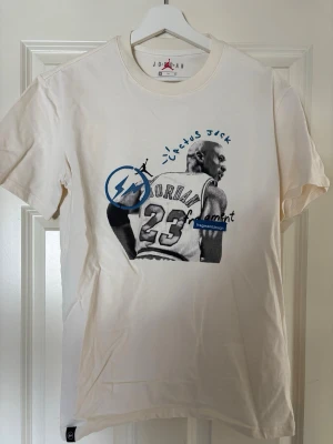 Jordan x Cactus Jack vit t-shirt - Vit t-shirt från Jordan x Cactus Jack med tryck av Michael Jordan på ryggen och blå detaljer. T-shirten har rund hals och korta ärmar. Materialet är mjuk bomull och passformen är normal. Perfekt för dig som gillar streetwear och basket.