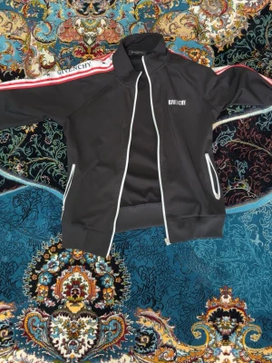 Svart track jacket från  - Det står fel storlek på tröjan men passar S/M. Snygg svart track jacket från med vita detaljer och dragkedja framtill. Röd och vit rand. Två fickor med vita kanter och hög krage för sportig look. 