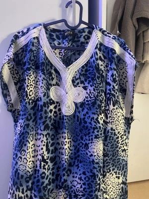Marockans gandoura - Säljer en blå och svart leopardmönstrad klänning med v-ringning och dekorativa vita detaljer vid halsen och axlarna. Klänningen har korta ärmar och ett avslappnat, loose fit. Perfekt för dig som gillar djurmönster och vill sticka ut. FRÅN MAROCKO