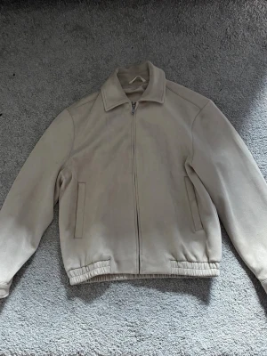 Beige bomberjacka från Zara - Stilren beige bomberjacka med dragkedja framtill och två sidofickor. Jackan har klassisk krage, resår vid midjan och långa ärmar. Perfekt för dig som gillar en clean och enkel look.