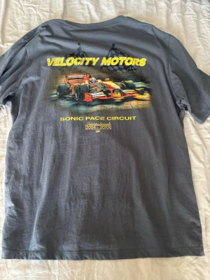 Grå Velocity Motors t-shirt - Den är grå det är skuggan som gör mönstret.Cool grå t-shirt med stort färgglatt tryck på ryggen av en formelbil och texten 'Velocity Motors' och 'Sonic Pace Circuit'. Perfekt för dig som gillar racing och vill sticka ut med en unik design. Skön passform och mjukt material.