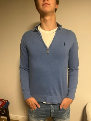 Blå halvziptröja från Ralph Lauren - Snygg blå stickad tröja med halv dragkedja från Ralph Lauren. Tröjan har ribbade muddar och krage, samt den klassiska broderade loggan på bröstet. Perfekt att bära över en t-shirt för en avslappnad men stilren look. Jävligt snygg men tyvärr blivit feltvättad, därav att den sitter lite smått. Modellen på bilden är 182 och väger 75kg. Storlek M men sitter som S/XS