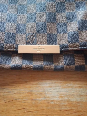 L*v väska  - Snygg axelväska från l*v i klassiskt brunt och mörkblått Damier-mönster. Väskan har en kombinerad axelrem i läder och kedja samt en metallplatta framtill med logotyp. Perfekt för dig som gillar lyxiga accessoarer.