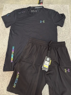 Svart träningsset från Under Armour - Snyggt svart träningsset från Under Armour med t-shirt och shorts. Setet har regnbågsfärgade loggor och textdetaljer på både tröja och shorts. Materialet är lätt och andas, perfekt för gymmet eller löprundan. Elastisk midja på shortsen för skön passform.