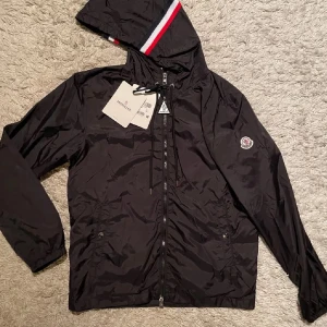 Svart vindjacka från Moncler med huva - Svart vindjacka från Moncler med huva och dragkedja framtill. Jackan har Moncler-logga på ärmen och snygga snören vid huvan. Röd, vit och blå randig detalj på huvkanten ger en sportig vibe. Perfekt för dig som gillar stilrena och exklusiva plagg. Taggen följer ej med