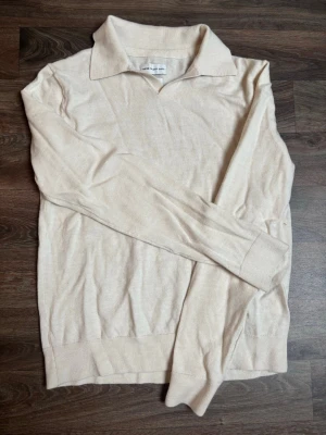 Beige merinoull tröja från these glory days - Säljer en stilren beige stickad tröja med klassisk krage och långa ärmar. Tröjan har ribbade muddar vid ärmslut och nederkant. Perfekt för dig som gillar en enkel men snygg look. Nypris 1000kr