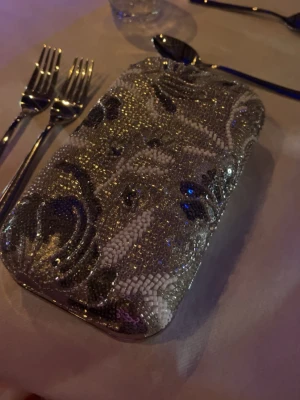 Glittrig silver kuvertväska - Säljer en glittrig kuvertväska i silver med mönster av små pärlor och paljetter i vitt, svart och grått. Väskan har en rektangulär form med rundade hörn och hårt skal. Perfekt accessoar för student balen!
