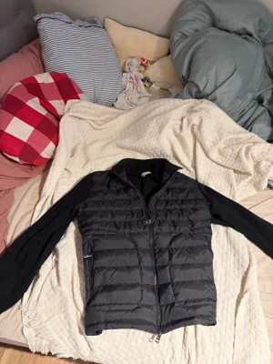 Svart cardiganjacka med dunväst - Snygg svart cardiganjacka från Moncler med vadderad dunväst framtill och stickade ärmar. Jackan har dragkedja, ståkrage och diskret Moncler-logga på ena ärmen. Perfekt för lager-på-lager och en stilren look.