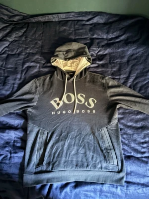 Hugo Boss hoodie  - Mörkblå hoodie från Hugo Boss med stor vit logga på bröstet och kontrastfärgad insida i huvan. Perfekt för en avslappnad och trendig look.