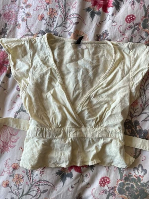 Ljusgul topp med knytband - Söt ljusgul topp med korta ärmar och knytband i midjan. Toppen har v-ringning och volangkant nedtill, vilket ger en feminin och avslappnad look. Perfekt till jeans eller kjol för en fräsch sommarstil.