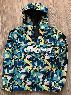 Ellesse camo vindjacka med huva - Färgstark vindjacka från Ellesse med blå, gul, svart och vit camo-mönster. Stor ficka framtill med Ellesse-logga, halv dragkedja och meshdetalj vid halsen. Jackan har huva och elastiska muddar. Perfekt för dig som vill sticka ut.