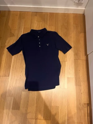 Mörkblå pikétröja från gant - Snygg mörkblå pikétröja från gant med klassisk krage och knappar framtill. Tröjan har korta ärmar och en liten broderad logga på bröstet. Perfekt till jeans eller shorts för en clean och stilren look.