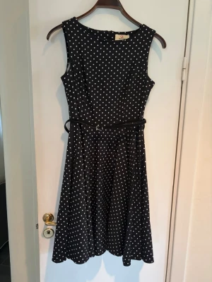 Svart prickig klänning 50-tal - Snygg svart klänning med vita prickar och ett smalt bälte i midjan i sammet. Ärmlös modell med rund hals och utställd kjol som ger en klassisk retrovibe. Perfekt för dig som gillar tidlös stil och vill sticka ut lite extra. Passar mig som en typisk 38-40 