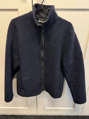 J Lindeberg fleecejacka - Mysig mörkblå fleecejacka med hel dragkedja framtill och hög krage. Jackan har långa ärmar och diskret logga på ena ärmen. Perfekt för kyliga dagar och enkel att matcha med olika outfits.