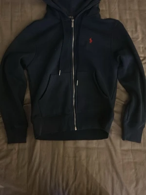 Mörkblå zip-hoodie från Polo Ralph Lauren - Stilren mörk blå hoodie med dragkedja från Polo Ralph Lauren. Klassisk huva med dragsko, två fickor framtill och den ikoniska röda loggan broderad på bröstet. Perfekt för en avslappnad och trendig look.
