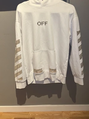 Vit Off-White hoodie med tryck - Säljer en vit hoodie från Off-White med silverfärgade detaljer. Framsidan har texten 'OFF' och ärmarna har de klassiska diagonala Off-White-ränderna. Baksidan pryds av det ikoniska Off-White-korset och texten 'SEEING THINGS'. Stor ficka framtill och huva.