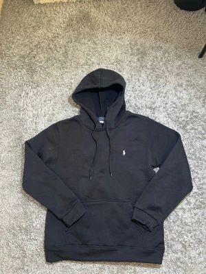 Svart hoodie från Polo Ralph Lauren - Svart hoodie med huva och dragsko från Polo Ralph Lauren. Klassisk design med broderad logga på bröstet och känguruficka framtill. Mjuk bomullskänsla och ribbade muddar vid ärmslut och nederkant.