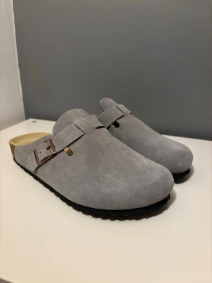 Gråa Birkenstock Boston - Säljer ett par gråa Birkenstock i mocka med justerbart metallspänne och svart räfflad sula. Tofflorna har en klassisk slip-on design och en bekväm innersula i kork. Perfekta för chill dagar hemma eller när du vill ha något enkelt och stilrent på fötterna. Modellen är Boston. Återkom vid frågor 😊
