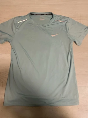 Ljusblå Nike miler tshirt - Snygg ljusblå tränings-tshirt från Nike med rund halsringning och korta ärmar. T-shirten har vita detaljer på axlarna och en vit Nike-logga på bröstet. Perfekt för löpning eller gymmet, tillverkad i ett lätt och snabbtorkande material.