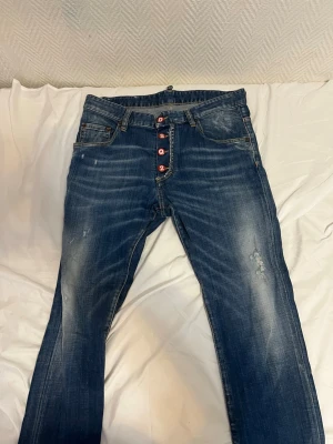 Dsquared2 jeans  - Säljer ett par blå jeans med snyggt slitna detaljer och röda knappar i gylfen. Jeansen har klassisk femficksmodell och raka ben. Materialet är denim i bomull och passformen är avslappnad men ändå stilren.