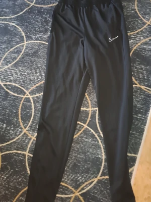 Svarta mjukisbyxor från Nike - Säljer ett par svarta mjukisbyxor från Nike med klassisk vit swoosh-logga på benet. Byxorna har resår i midjan och är tillverkade i ett mjukt och stretchigt material som passar perfekt för chill eller träning. Snygg och enkel design som funkar till det mesta.