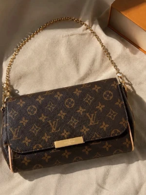 Damväska  - Snygg handväska i klassiskt brunt monogrammönster med guldiga detaljer och kedjehandtag. Väskan har en rektangulär form och stängs med lock. Perfekt accessoar för dig som gillar lyxig och tidlös design.