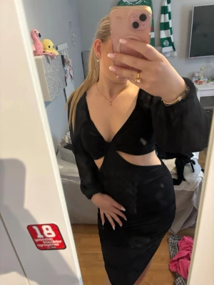 Svart långärmad cut out klänning - Snygg svart klänning med lång ärm och djup v-ringning. Klänningen har trendiga cut out-detaljer vid midjan och twistad front. Materialet ser ut att vara mjukt och följsamt, perfekt för en stilren look.