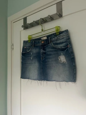 Blå jeanskjol med slitningar - Säljer en kort blå jeanskjol med rå kant och slitna detaljer. Kjolen har klassisk femficksdesign, knapp och dragkedja framtill samt en snygg tvättad look. Perfekt för dig som gillar en avslappnad och trendig stil.