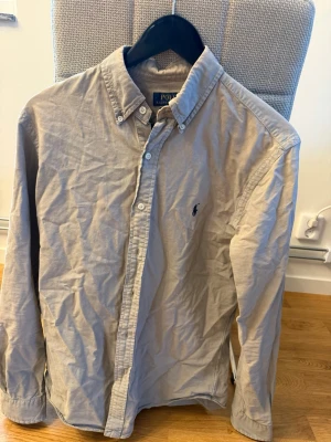Beige skjorta från Polo Ralph Lauren - Klassisk beige skjorta från Polo Ralph Lauren i slim fit-modell. Skjortan har långa ärmar, knappar framtill och den ikoniska broderade loggan på bröstet. Tillverkad i mjuk bomull och passar perfekt till en avslappnad men ändå stilren look.