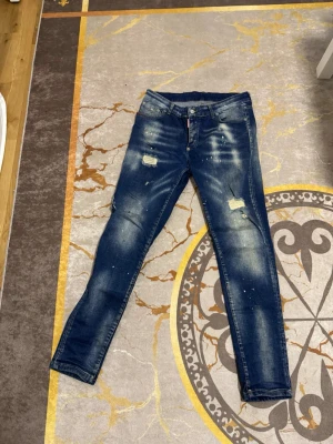Mörkblå slitna skinny jeans från Dsquared2 - Säljer ett par mörkblå skinny jeans med slitningar och distressed detaljer. Jeansen har ljusa tvättade partier, färgstänk och klassisk femficksmodell. Perfekta för en trendig och avslappnad stil.